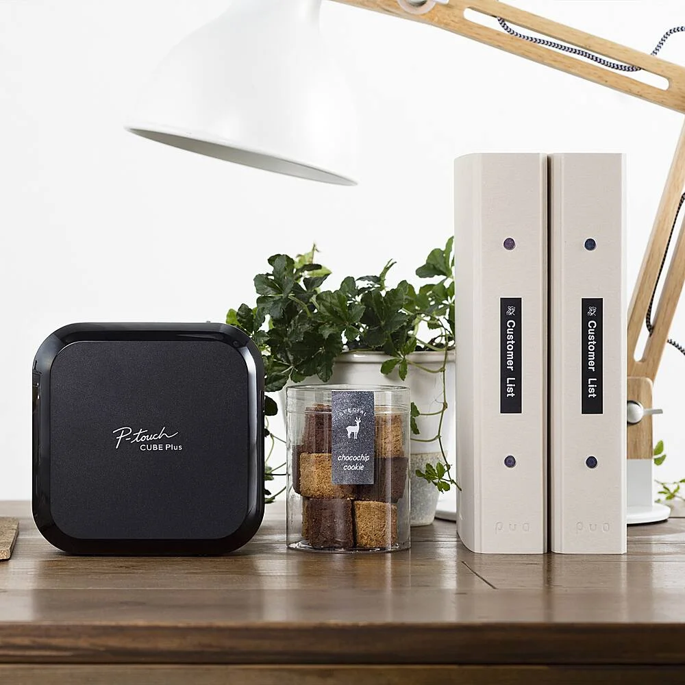Brother P-Touch Cube Plus PT-P710BT on a desk with labeled binders and jar showing printed label examples