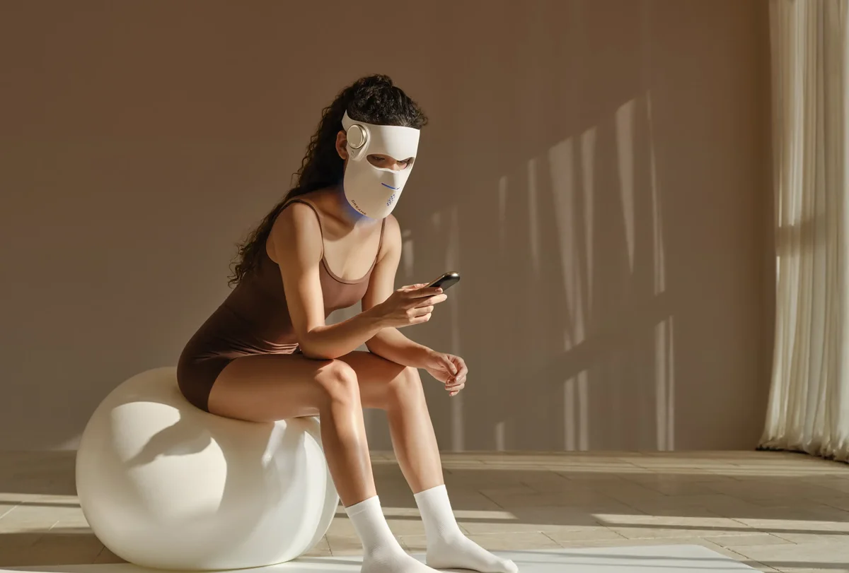 Woman wearing the Dreame Chrona LED mask with a blue glow at the chin, sitting on an exercise ball and using the wireless remote in a sunlit room