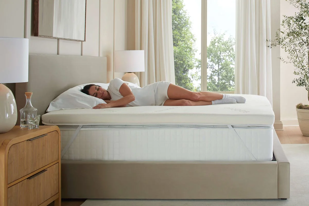 Saatva Graphite Memory Foam Mattress Topper installed on a bed with a woman resting on the surface, showing the elastic corner straps in use