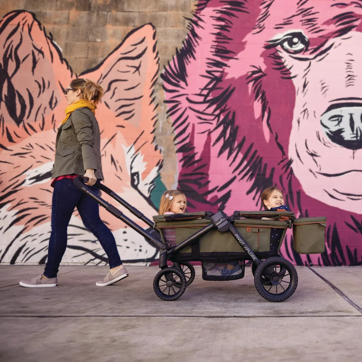 Mom pulling two children in Evenflo Pivot Xplore wagon in pull mode past a colorful mural