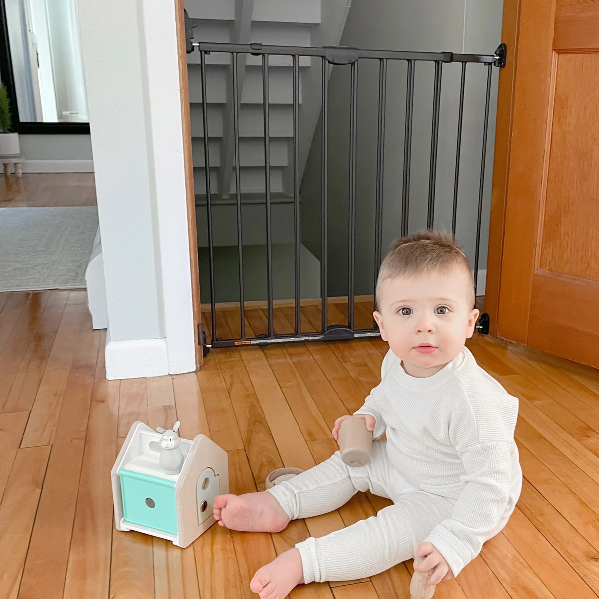 Baby sitting on hardwood floor playing with toys in front of the closed Toddleroo baby gate at base of stairs