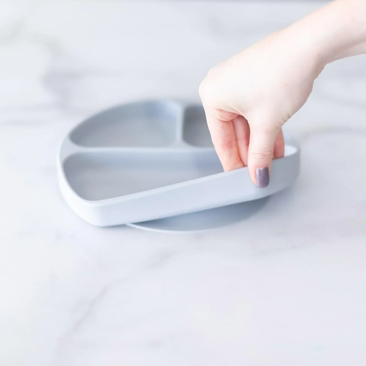 Hand lifting the Bumkins Silicone Grip Dish to show the suction base releasing from a marble surface