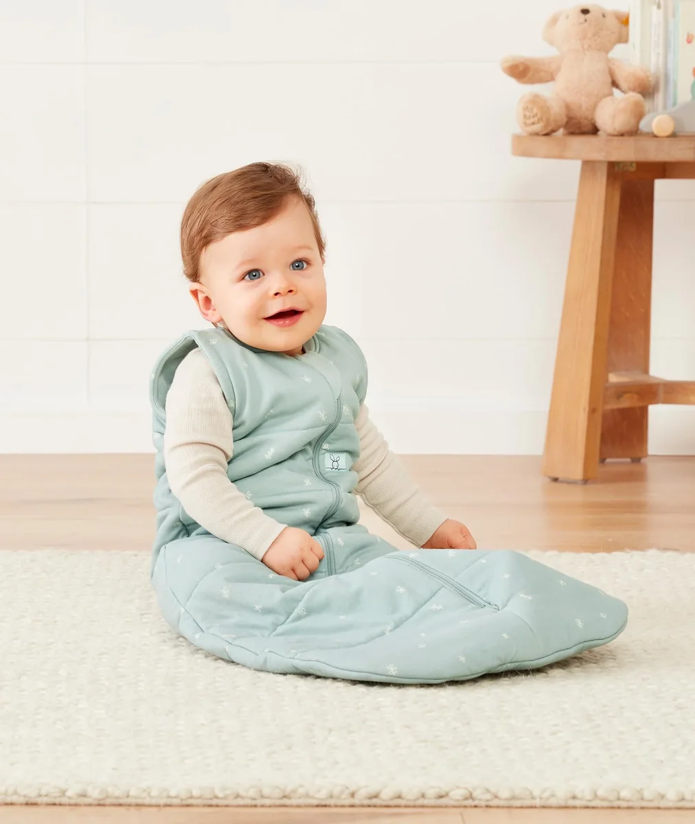 Baby sitting on a rug wearing the sage ergoPouch Sleep Sack over a long-sleeve top