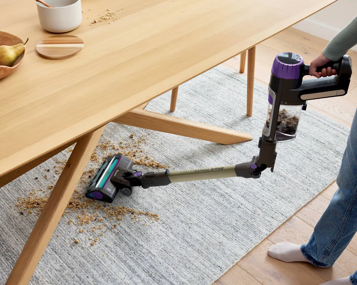 Shark PowerDetect Clean & Empty with its MultiFlex wand bent to reach under a dining table, the DuoClean Detect floor head picking up spilled cereal from a rug