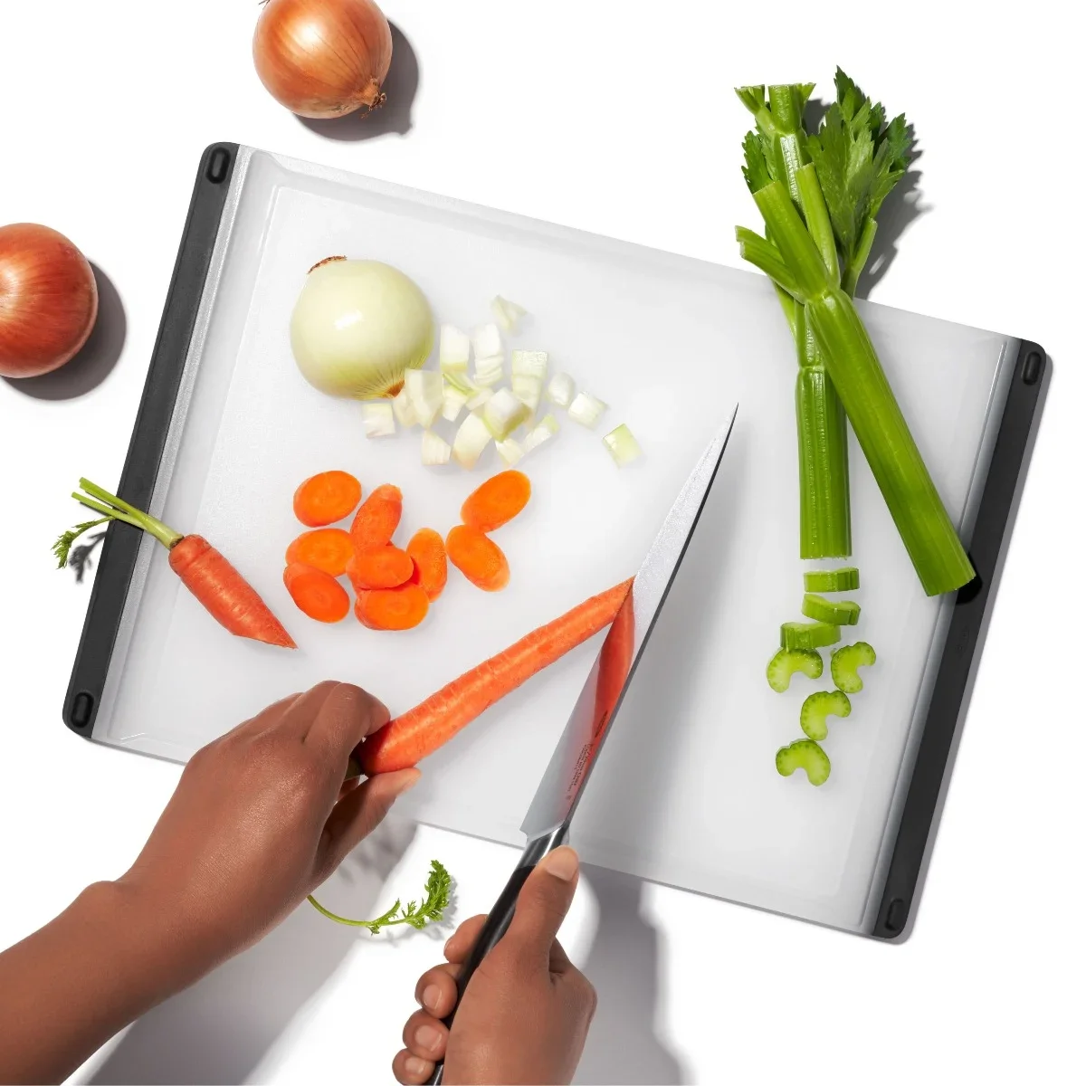 Hands using a knife to chop carrots, onion, and celery on the OXO Good Grips cutting board, showing the full board in use with its non-slip feet