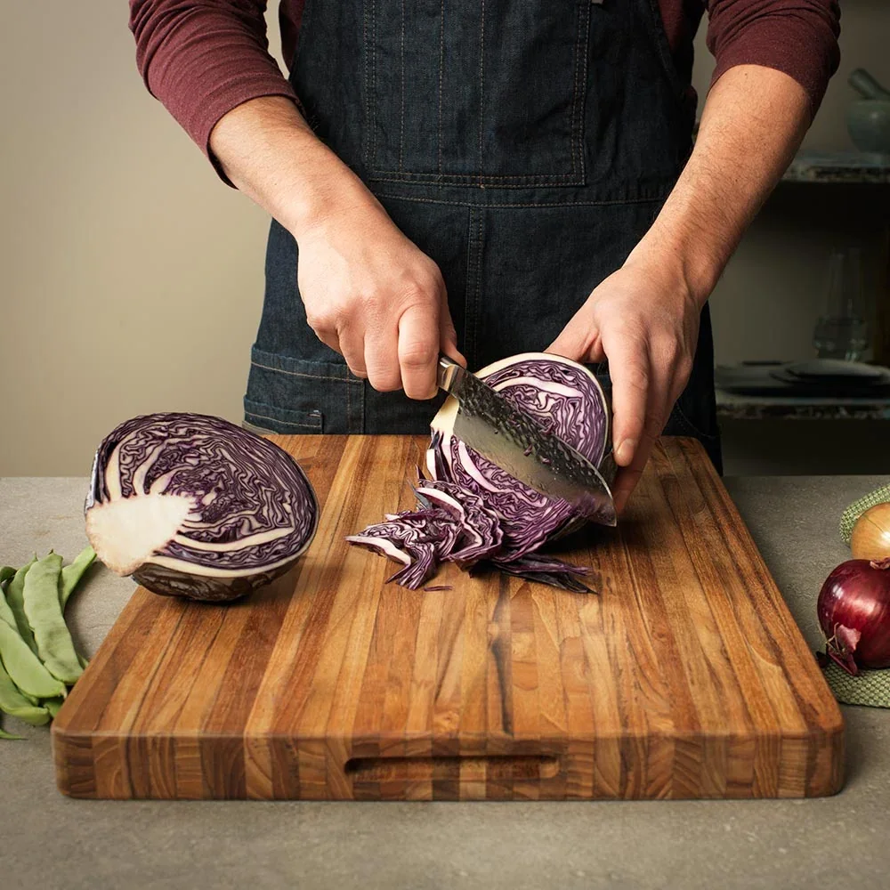 A cook in an apron slicing red cabbage on the Teakhaus Extra-Large Professional Cutting Board, with green beans and a red onion nearby on the counter