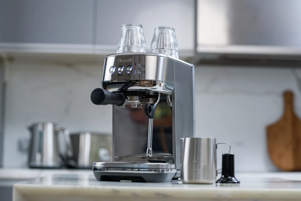 Breville Bambino Plus on a marble kitchen counter with a milk jug, tamper and glasses warming on top