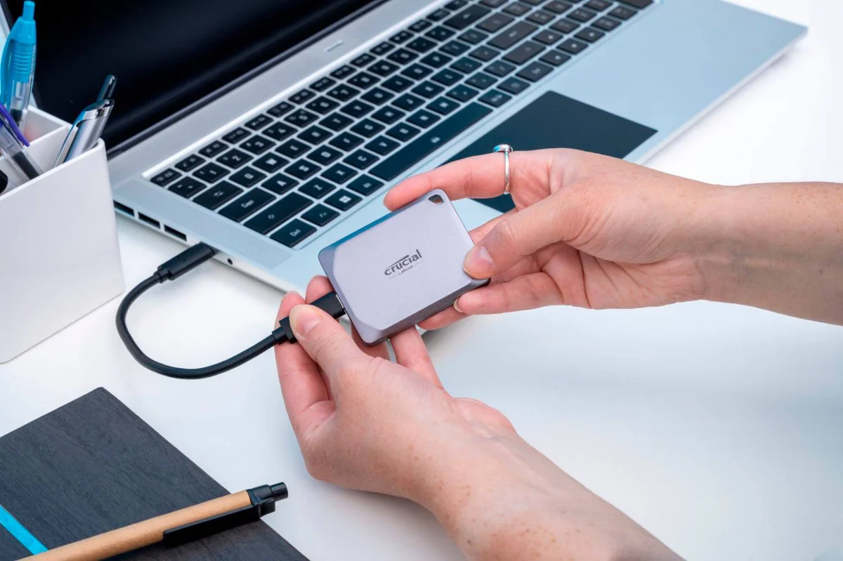 Crucial X9 Pro portable SSD held in hand and plugged into a laptop via USB-C, showing its tiny credit-card-like scale