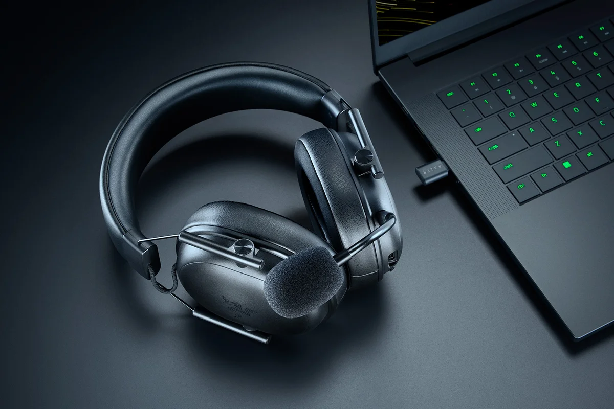 Razer BlackShark V3 X HyperSpeed lying on a desk beside a laptop with its 2.4GHz HyperSpeed USB dongle plugged in