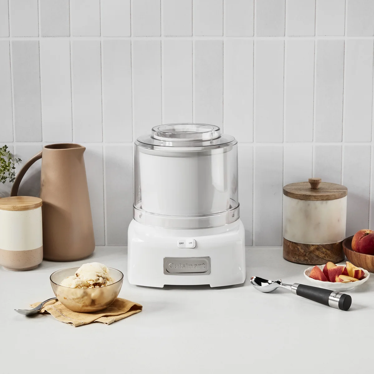 Cuisinart ICE-21P1 ice cream maker in use on a kitchen counter with a bowl of finished ice cream, waffle cones, and fresh peaches