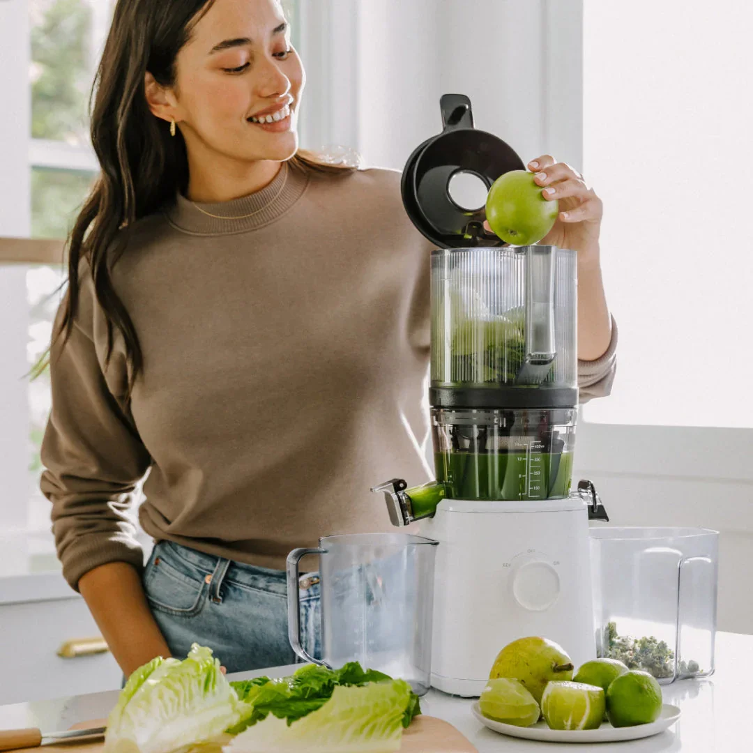 Person loading a whole apple into the Nama J2 Cold Press Juicer hopper with green juice and produce on counter