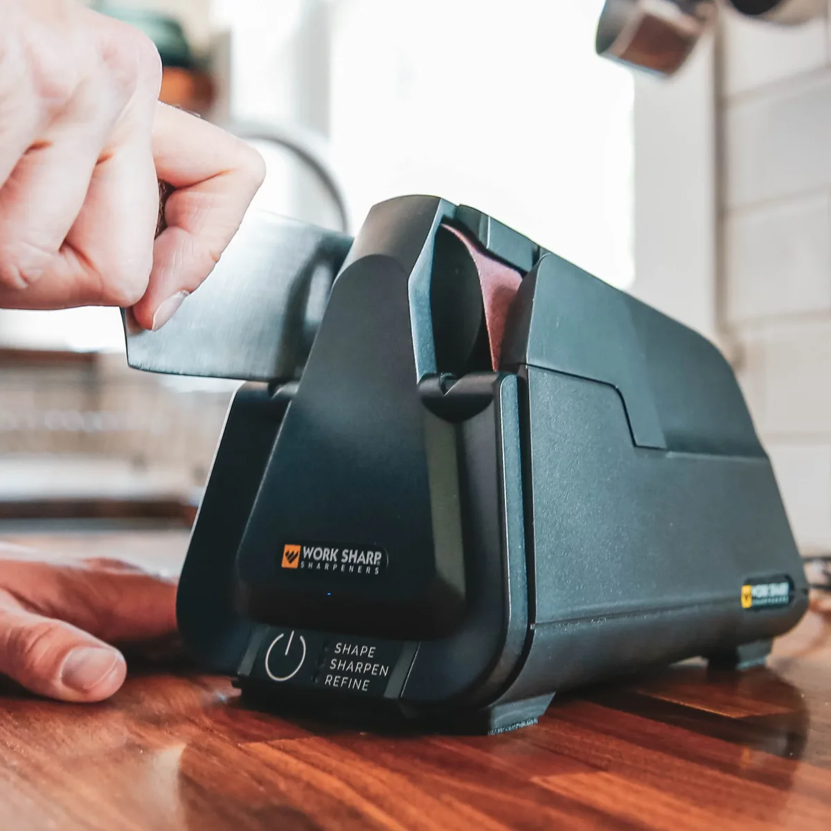 Close-up of a hand guiding a knife blade into the Work Sharp Professional Electric Kitchen Knife Sharpener's belt slot on a wooden kitchen counter