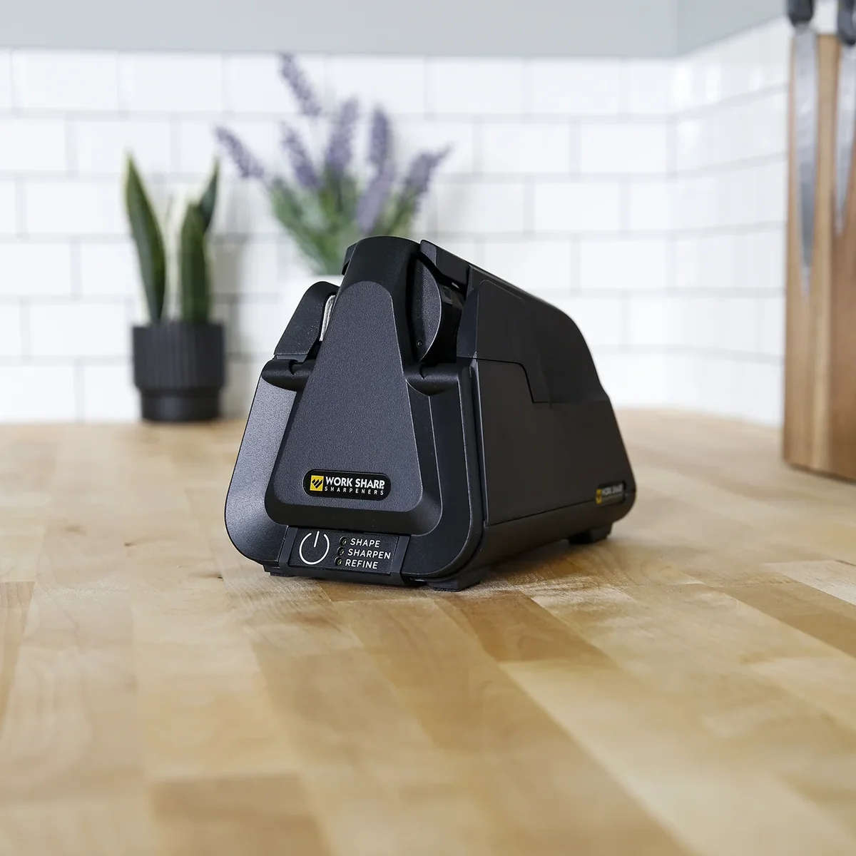 Work Sharp Professional Electric Kitchen Knife Sharpener sitting on a butcher block kitchen counter with a knife block and plants in the background