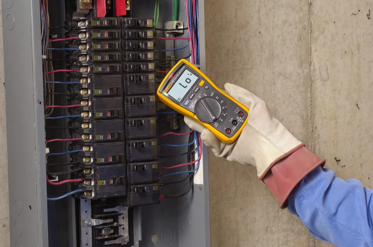 Fluke 117 multimeter in use, held in a gloved hand while testing wiring inside an electrical breaker panel