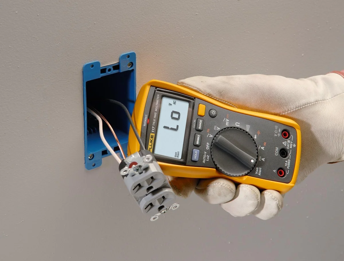 Fluke 117 multimeter in use, held in a gloved hand checking voltage at an outlet in a wall box