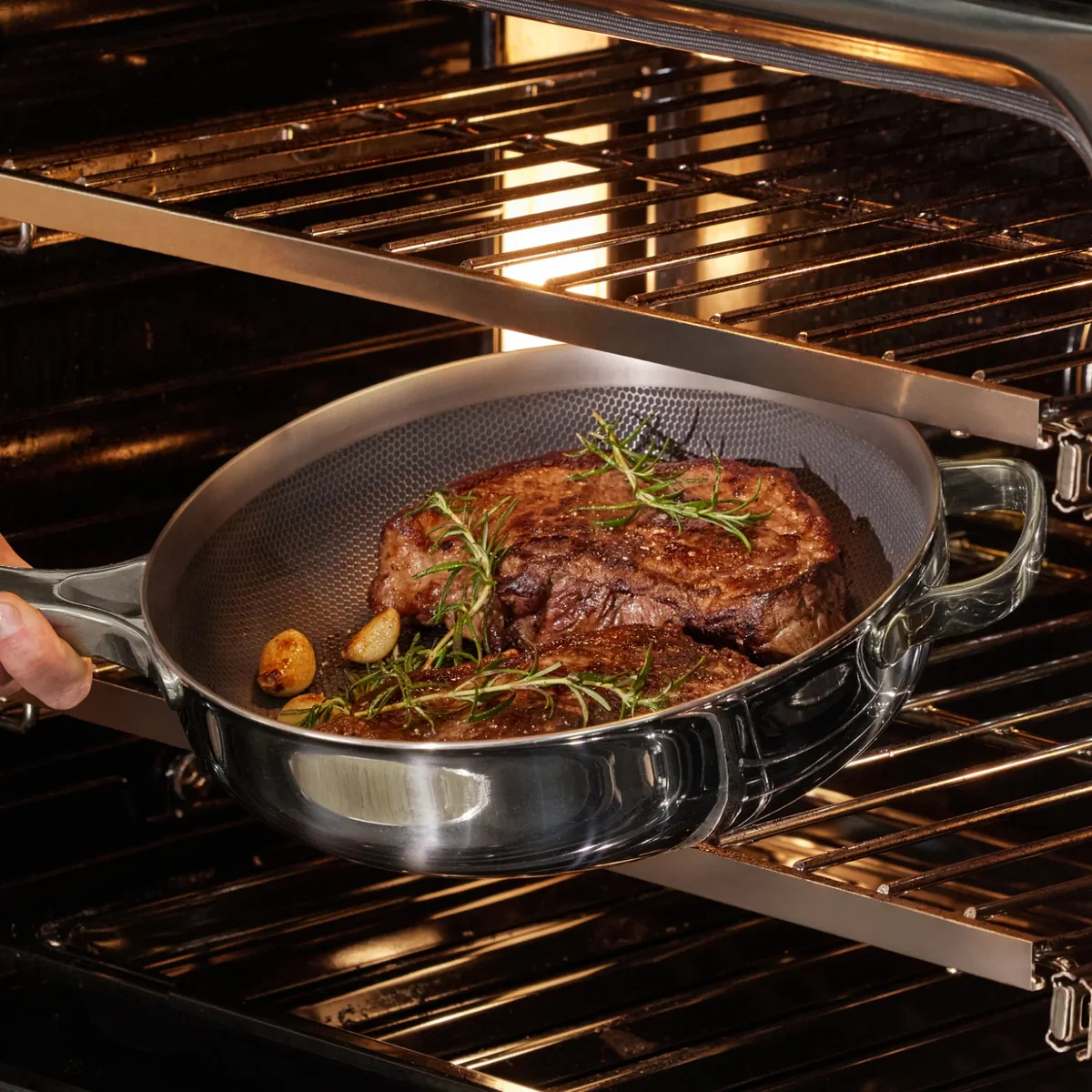 Our Place Titanium Always Pan Pro holding a seared steak with rosemary and garlic inside a home oven, showing the honeycomb-textured titanium interior