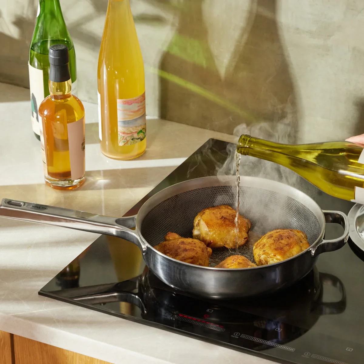 Our Place Titanium Always Pan Pro searing chicken thighs on an induction cooktop with wine being poured in, showing the mirror-polished chrome exterior