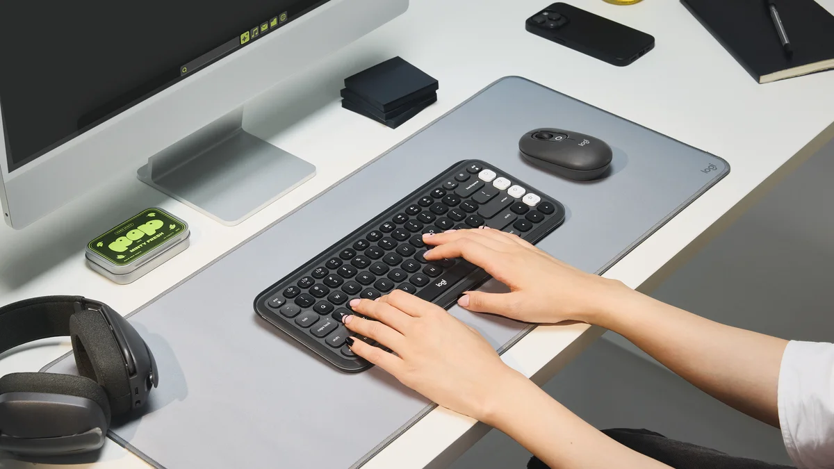 Hands typing on the Logitech POP Icon Keys on a desk mat next to a matching POP Mouse and a monitor