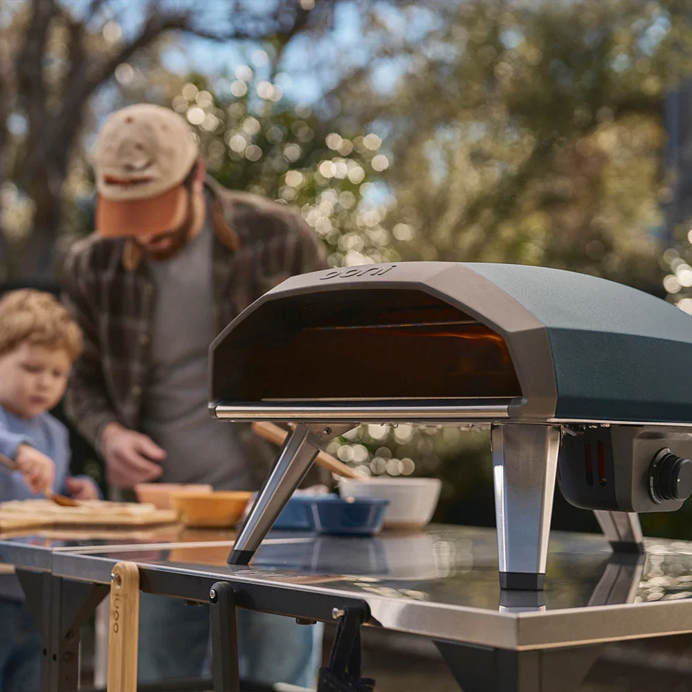 Ooni Koda 2 in slate blue set up on an outdoor table with a family in the background and a bamboo pizza peel resting against the stand