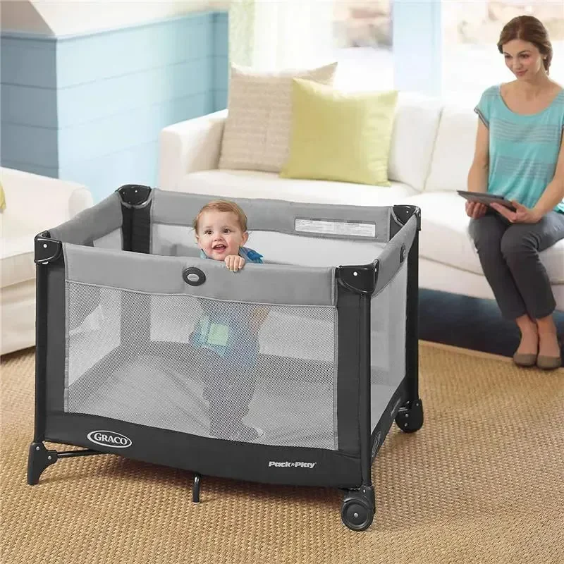 Graco Pack 'n Play playard in living room with toddler standing inside and mother relaxing nearby