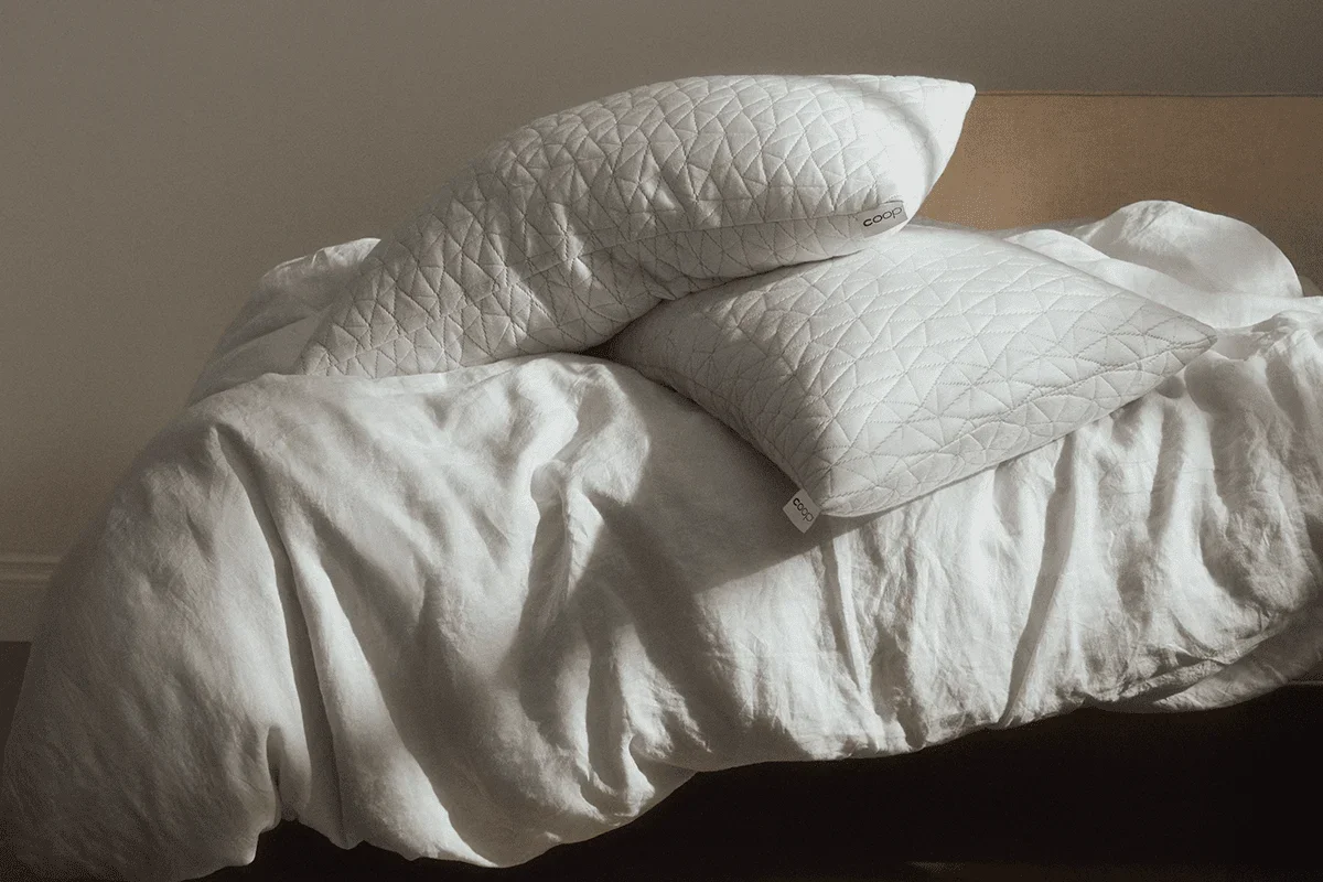 Two Coop Original Adjustable Pillows resting on a bed with rumpled white linen bedding