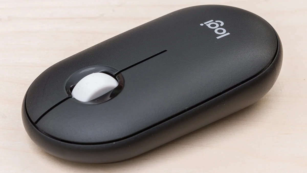 Logitech Pebble Mouse 2 M350s in graphite, top-down 3/4 view on light wood surface