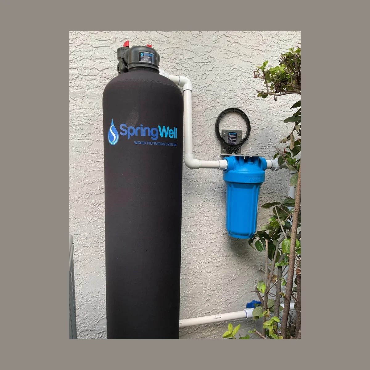 SpringWell CF1 carbon filter tank and blue sediment pre-filter housing installed outdoors against a stucco wall