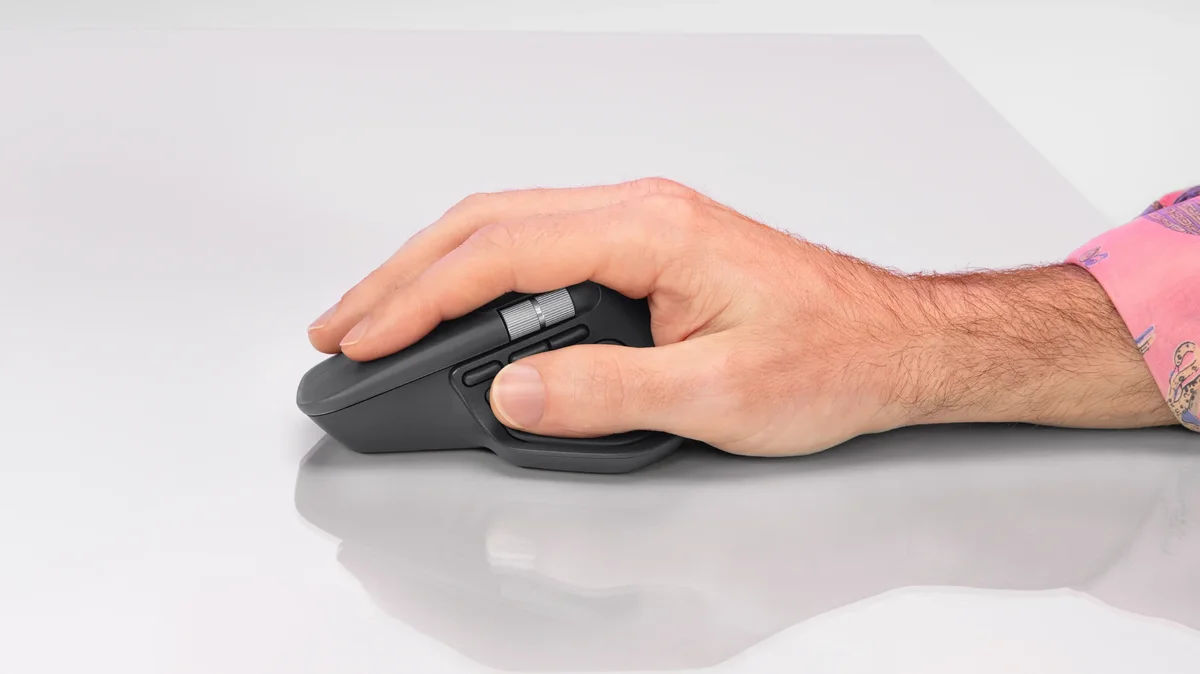 Hand gripping the Logitech MX Master 4 on a light desk, side view with the thumb resting on the haptic sense panel below the thumb wheel